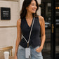 A woman wearing a black sleeveless vest with a pearl-bead crossbody strap and a white phone case, standing on a city sidewalk next to a cafe table.